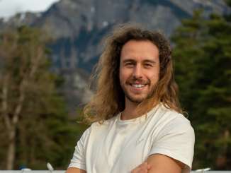 Giovane uomo con lunghi capelli castani sorride alla telecamera