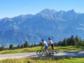 Due persone scendono in scooter alpino e sullo sfondo si intravedono le montagne della Bündner Herrschaft