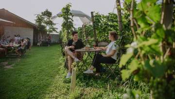 Due persone siedono tra le viti e si godono un bicchiere di vino insieme.